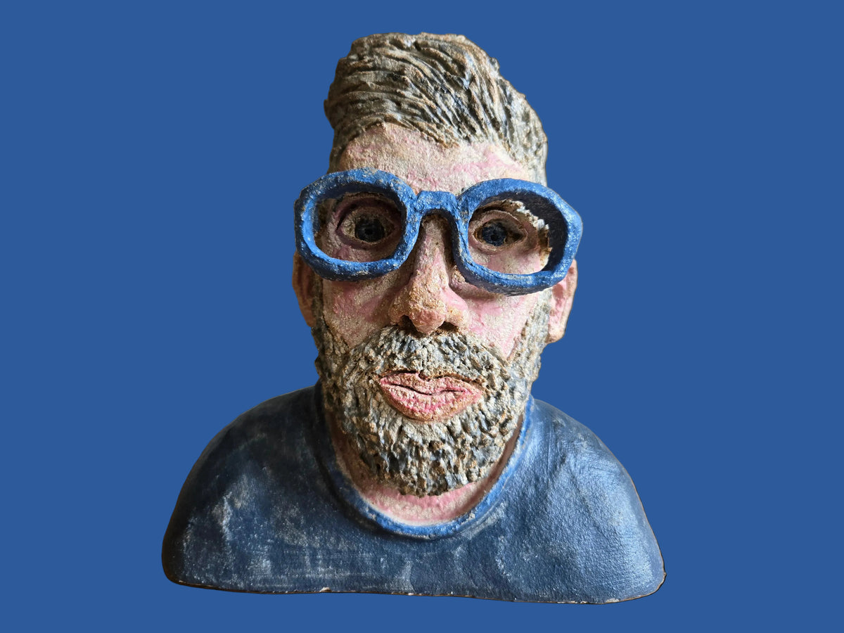 A handmade clay or ceramic bust sculpture of a man with a beard, wearing blue-rimmed glasses and a blue shirt.