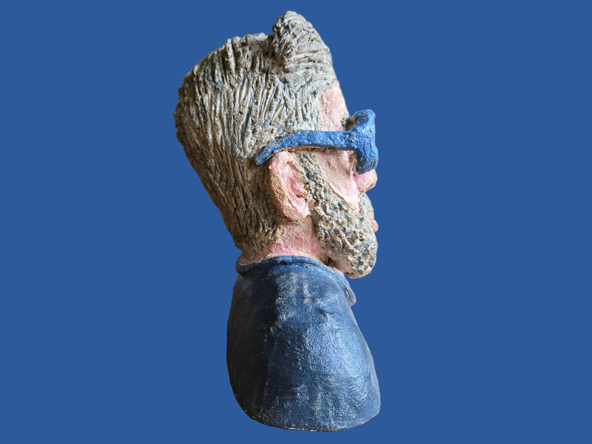 A sculptural bust of a man's head in profile, featuring textured grey hair, a grey beard, pinkish skin tones, blue eyepieces, and a dark blue shirt.