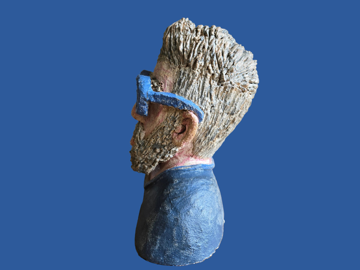A sculptural bust of a man's head in profile, featuring textured grey hair and beard with blue glasses and a blue shirt.